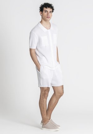 Joven de pie con las manos en los bolsillos, vistiendo una camiseta polo de punto blanca de manga corta, pantalones cortos a juego de punto y zapatillas beige sobre fondo blanco.