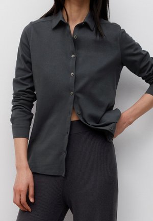 Femme portant une chemise à manches longues boutonnée gris foncé partiellement rentrée dans un pantalon taille haute côtelé gris foncé, debout devant un fond uni.