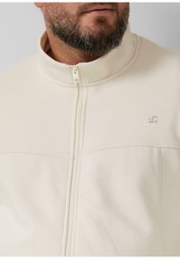 Veste zippée beige clair avec un col montant, tissu texturé et détail de logo subtil sur la poitrine. Fini lisse sans motifs visibles.