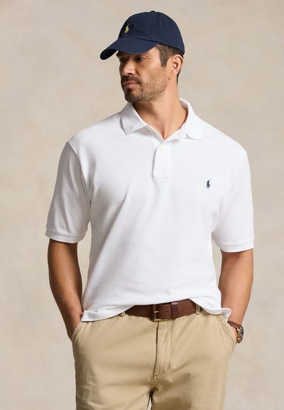 Mann, der ein weißes Polohemd, beige Hose, braunen Gürtel, Uhr und eine marineblaue Baseballkappe trägt, steht mit den Händen in den Taschen und blickt zur Seite.