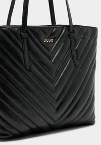 Zwarte gewatteerde shopper in kunstleer, met een chevronpatroon, dubbele handvatten en metalen logodetail. Gladde textuur met een gestructureerde vorm.