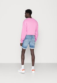 Felpa corta rosa, pantaloni corti di jeans azzurri con risvolti, sneakers bianche con dettagli arancioni e pelle scura. Sfondo semplice.