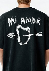 T-shirt en coton noir à coupe oversize, avec un graphique blanc au dos incluant le texte "Mi Amor" et une flèche traversant un cœur.