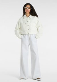 Geknipte witte denimjas met twee voorkruiken en zilveren knopen, gecombineerd met lichtgrijze wijduitlopende sweatpants en witte schoenen.