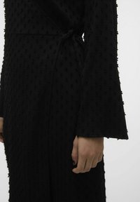 Zwarte jurk van textuurstof met verhoogde stippen, lange mouwen en een strik in de taille. Een mooie afwerking met een lichte glans is zichtbaar.