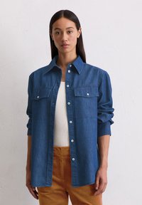 Blauwe denim overhemd met button-down kraag, twee voorzakken en lange mouwen, gedragen over een wit top en gecombineerd met oranje broeken.