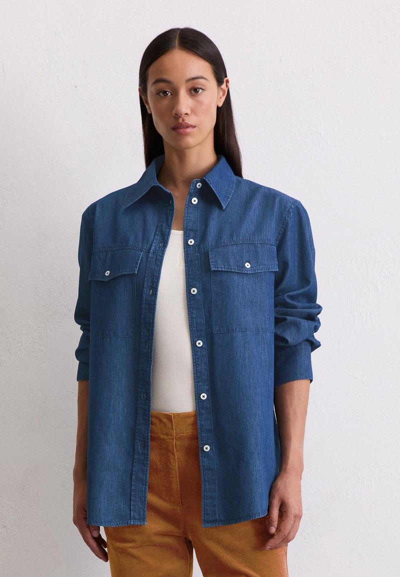 Blauwe denim overhemd met button-down kraag, twee voorzakken en lange mouwen, gedragen over een wit top en gecombineerd met oranje broeken.