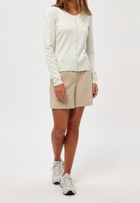 Ljusgrön cardigan med knappar framtill, ribbade muddar och en ribbad nederkant. Kombination med beiga shorts och grå sportskor. Slät textur.