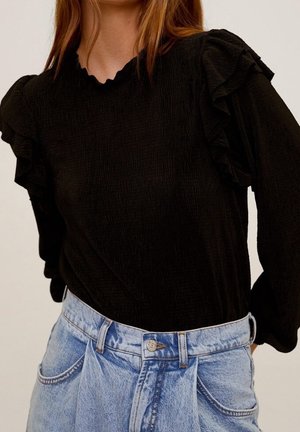 Kobieta ubrana w czarną bluzkę z długim rękawem z marszczonymi ramionami, włożoną w jasnoniebieskie dżinsy z wysokim stanem.