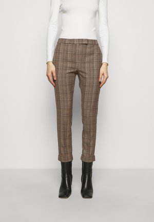 Femme portant un pantalon marron à carreaux taille haute, ajusté, avec des ourlets retroussés et des bottines noires, associé à un haut blanc à manches longues.
