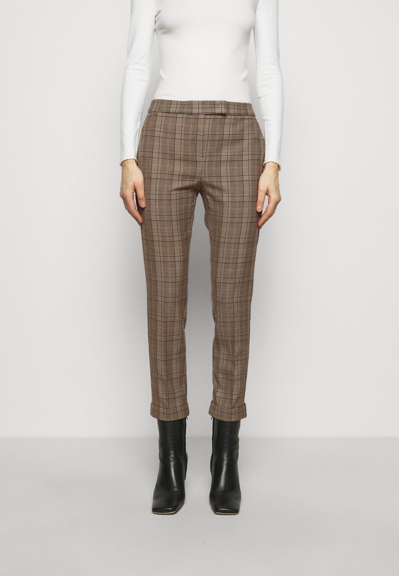 Femme portant un pantalon marron à carreaux taille haute, ajusté, avec des ourlets retroussés et des bottines noires, associé à un haut blanc à manches longues.
