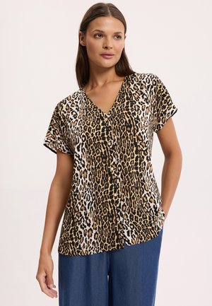 Kobieta ubrana w krótką koszulę na guziki w panterkę i niebieskie spodnie, stojąca z jedną ręką w kieszeni na jednolitym tle.