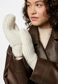 Creme strikkede vanter med en ribbet tekstur, der har et rum til tommelfingeren og fingrene, båret af en person i en brun læderjakke.