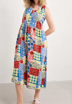 Kobieta ubrana w bezrękawnikową, wielokolorową sukienkę midi w stylu patchwork z kwiatowymi i liściastymi wzorami, do kompletu jasnoróżowe sandały na sprzączkę.