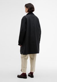 Casaco oversized preto com colarinho, punhos com botões e fendas laterais, combinado com calças bege e sapatos tipo loafers escuros, em fundo branco.