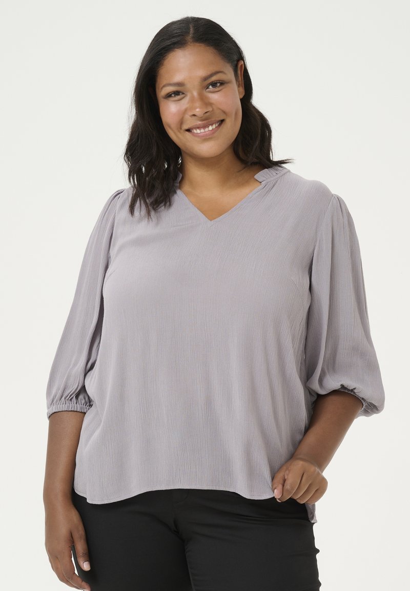 Blusa gris claro con escote en forma de V, mangas largas abullonadas y corte relajado. Tejido texturizado con sutiles líneas verticales y detalles de mangas fruncidas.