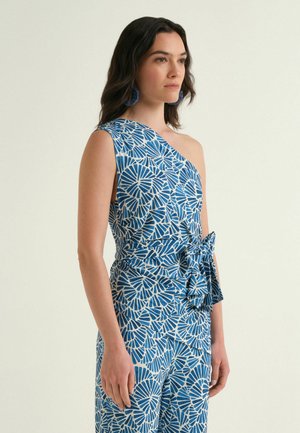 Femme portant une combinaison une épaule à motif bleu et blanc avec un nœud noué à la taille et des boucles d'oreilles bleues assorties, debout devant un fond uni.