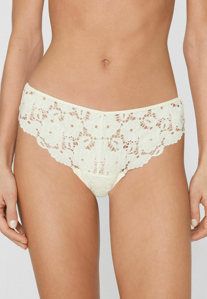 Woman wearing cream lace floral-patterned underwear, showing midsection and upper thighs against a plain background.