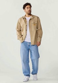 Beige skjorte med knapper og en lomme samt et mærke, båret over en hvid t-shirt; sat sammen med lyseblå baggy jeans og hvide sneakers.