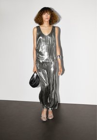 Metalické stříbrné top bez rukávů a široké kalhoty, doplněné sandály s průhlednými řemínky. Drží malou černou kabelku; hladká, lesklá textura.