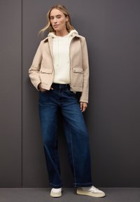 Beżowa wełniana kurtka z dwoma przednimi kieszeniami, połączona z kremowym hoodie i ciemnoniebieskimi spodniami o szerokich nogawkach, uzupełniona białymi trampkami.