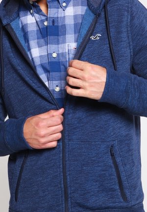 Man wearing a blue plaid button-up shirt and blue zip-up hoodie with pockets, holding the hoodie’s zipper near the chest.