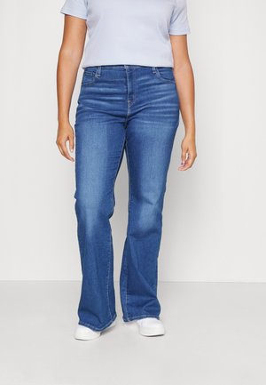 Person iført blå bootcut jeans, hvide sneakers og en lysefarget kortærmet skjorte, stående mod en neutral baggrund.