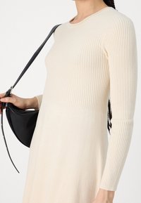 Abito a maglia a coste color crema con maniche lunghe, scollatura rotonda e orlo svasato, abbinato a una borsa curva nera. Tessuto morbido e design attillato.