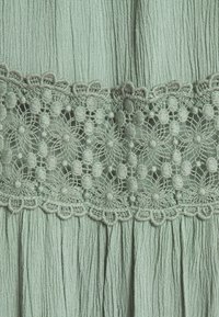 Tela verde claro con un acabado texturizado, que presenta un patrón decorativo de encaje a lo largo de la cintura, detallado con motivos florales y bordes en forma de ondas.