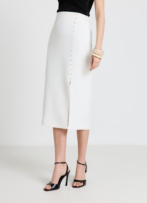 Donna che indossa una gonna midi bianca con bottoni frontali e spacco, abbinata a un top nero senza maniche, sandali neri con tacco alto e braccialetti beige.