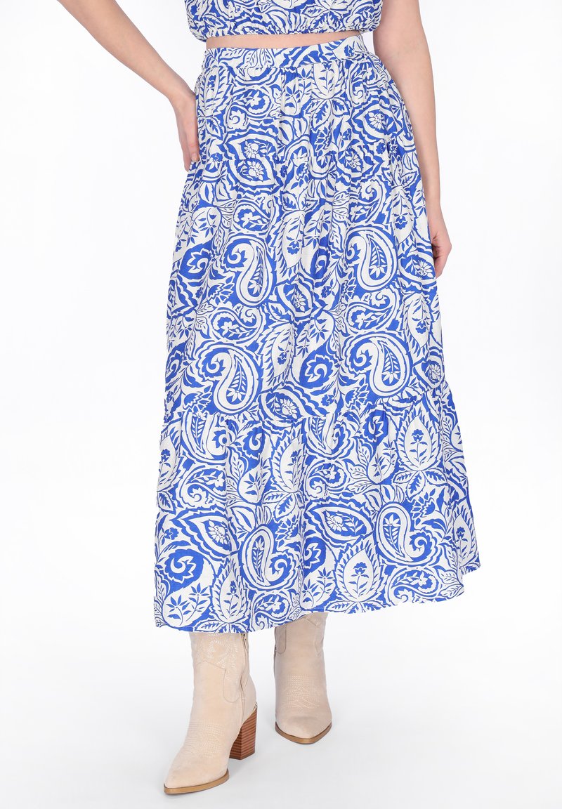 Blauer und weißer Rock mit Paisley-Muster, getragen mit beigen Knöchelstiefeln, Hand auf der Hüfte, weißer Hintergrund.