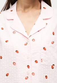 Chemise rose clair à boutons avec petits imprimés rouges de fraises et une poche poitrine à l'avant.