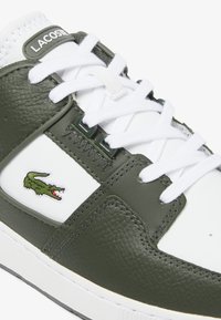 Sneaker verde e bianco Lacoste con pelle testurizzata, lacci bianchi e emblema del logo. Forma arrotondata con suola in gomma bianca e dettagli di cucitura.