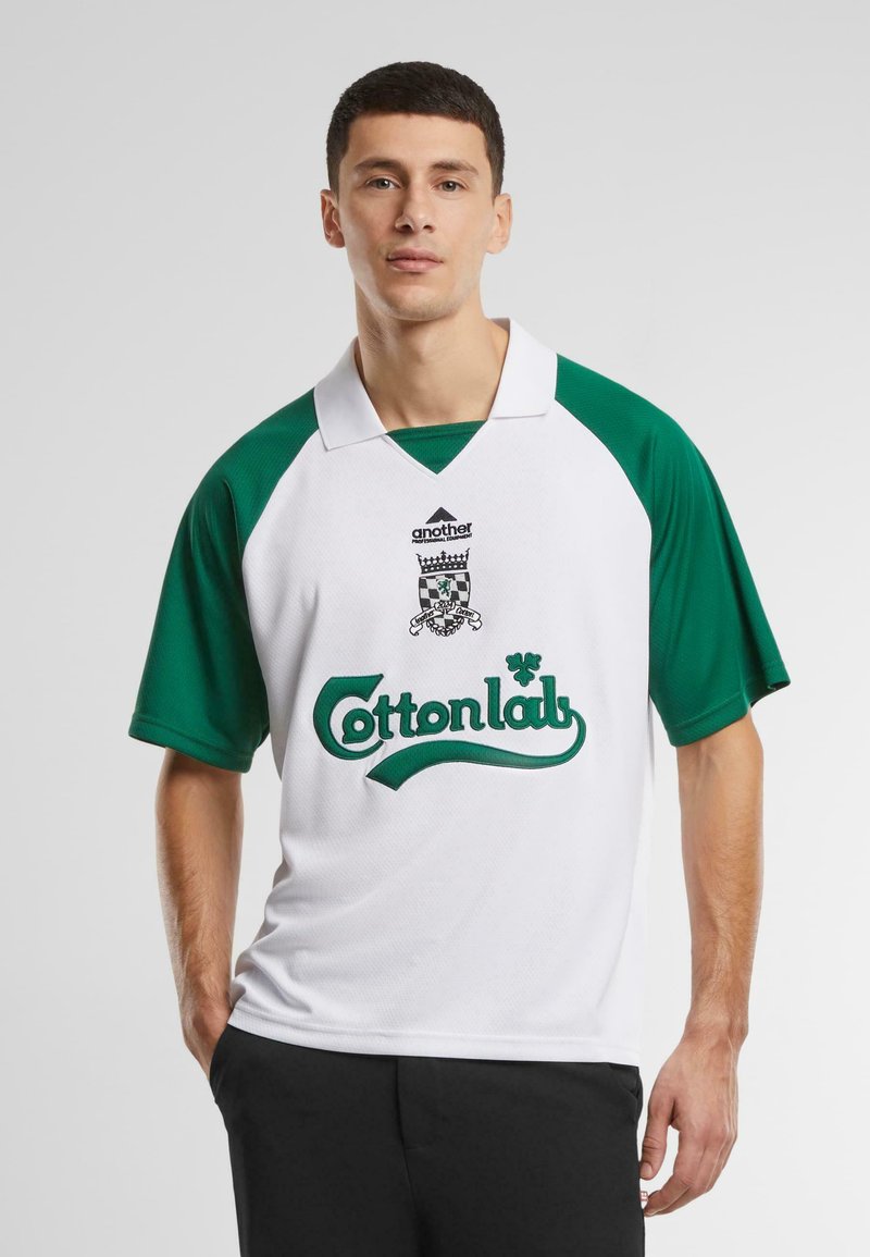 Hvit sportsdrakt med grønne raglanermer. Har en logo med teksten "Cottonlab" og et dekorativt emblem på brystet. Lett stoff.