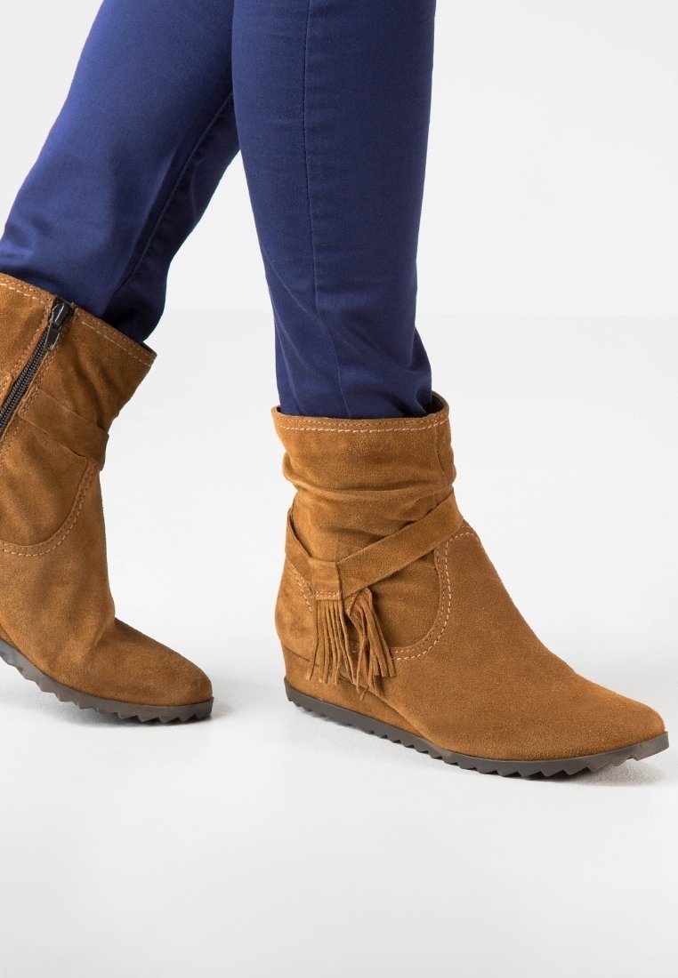 Bruine suède enkellaarsjes met een slouchy ontwerp, een zijrits en een kwastje. De zool is textuur voor grip. G gedragen met donkerblauwe broek.