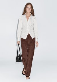 White long-sleeve button-up shirt knotted at the waist, paired with brown suede wide-leg pants, black handbag, and strappy black sandals.