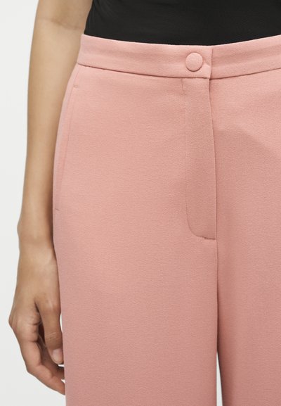 Γυναίκα που φοράει ψηλόμεσο, ανοιχτό ροζ σακάκι με κουμπί καλυμμένο με ύφασμα και πλαϊνές τσέπες, σε συνδυασμό με μαύρο τοπ.