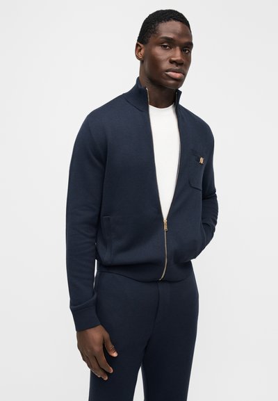 Marineblauwe sweater met rits, geribbelde breistructuur, col met opstaande kraag, gouden rits, borstzak met merklogo, gecombineerd met bijpassende marineblauwe broek.