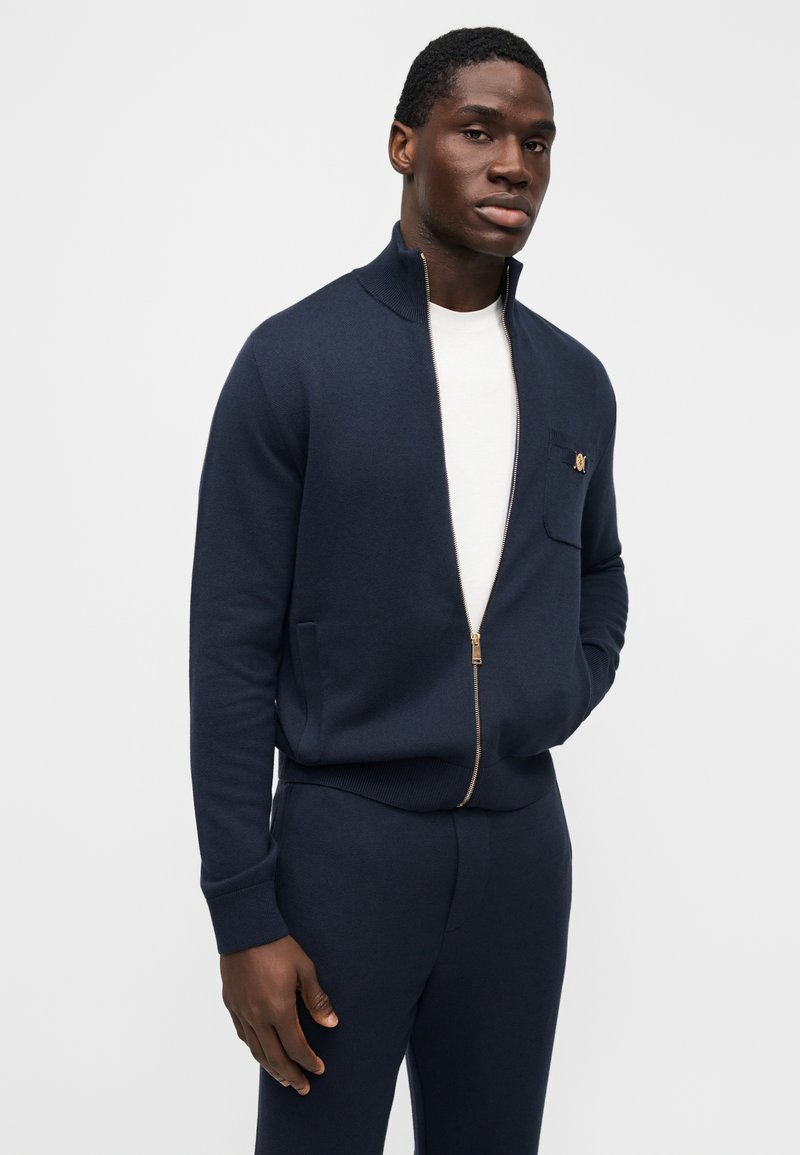 Marineblå zip-up-tröja med ribbad stickad textur, mockakrage, guldfärgad dragkedja, bröstficka med branding, matchande marinblå byxor.