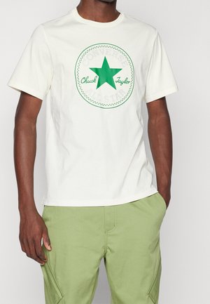 T-shirt en coton blanc avec un grand logo étoile verte accompagné du texte "Chuck Taylor", design circulaire avec une bordure cousue, porté avec un pantalon vert.