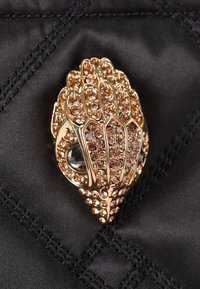 Spilla in tono oro a forma di mano, decorata con scintillanti pietre marrone chiaro, posta su uno sfondo di tessuto nero testurizzato.
