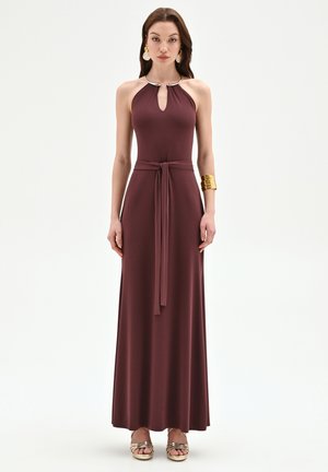 Femme portant une robe maxi bordeaux sans manches à col halter avec ceinture à la taille, bracelet manchette doré, boucles d'oreilles dorées et talons ouverts dorés.