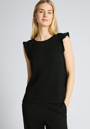 Femme blonde aux cheveux lisses portant un haut noir texturé sans manches et un pantalon noir, debout avec une main dans la poche.