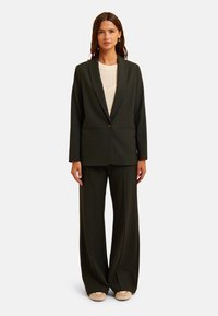 Blazer sartorial verde scuro con rever a punta e chiusura a un bottone, abbinato a pantaloni a gamba larga. Sotto, una maglietta leggera con un motivo testurizzato.