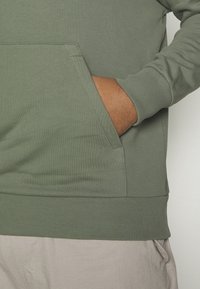 Felpa verde in cotone con tasca a marsupio frontale, polsini e orlo ribattuti, e una texture liscia. Dettagli delle cuciture visibili lungo la tasca.
