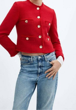 Mujer con una chaqueta roja ajustada de textura con botones dorados y vaqueros de cintura alta azul claro, con la mano apoyada en la cadera.