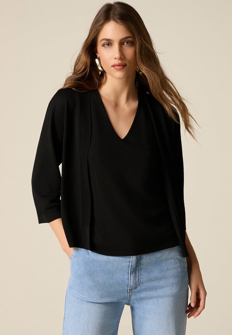 Donna con capelli lunghi e ondulati che indossa orecchini dorati, una maglietta a V nera con maniche a 3/4 e jeans azzurri, su sfondo beige chiaro.