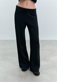 Sorte sweatpants med snoretræk i taljen, brede ben og synlige sømme. Materialet virker blødt med en glat tekstur. Båret med sorte sko.