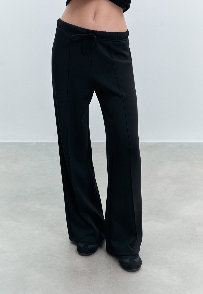 Sorte sweatpants med snoretræk i taljen, brede ben og synlige sømme. Materialet virker blødt med en glat tekstur. Båret med sorte sko.