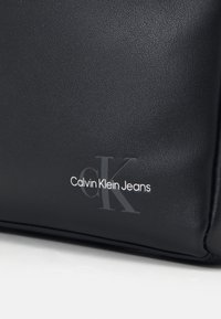 Bolso de mano negro texturizado hecho de material sintético que presenta el logo en relieve "Calvin Klein Jeans" en blanco, con un sutil acento de diseño "ck".